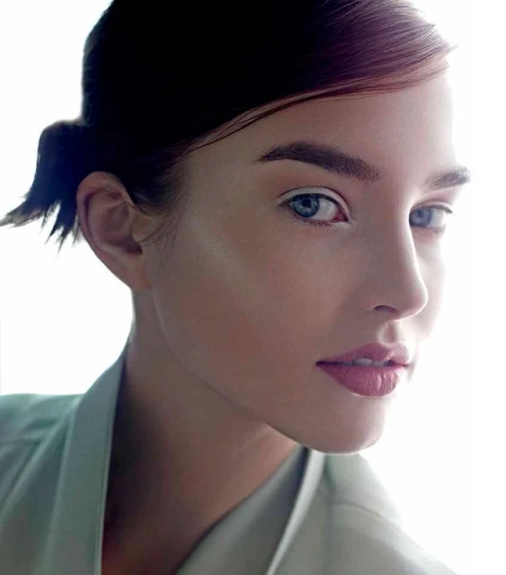 Close-up studio shot of a woman with light skin, blue eyes, and pink lips, wearing a light green jacket, against a bright white background.