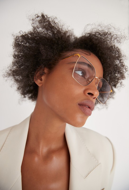 Close-up of an African American woman with afro hair wearing clear lens geometric gold-frame glasses and an off-white blazer in a studio.