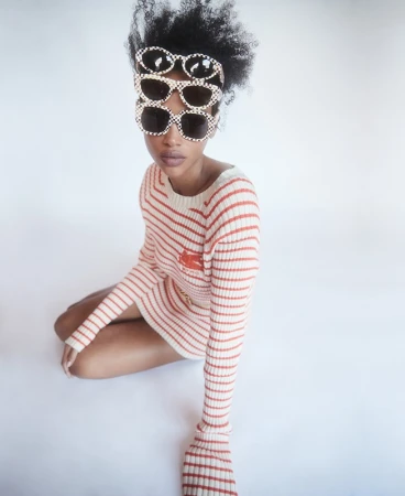 Stylish young woman sits with stacked sunglasses and a striped dress in a white studio setting, fashion portrait, eye-level angle.