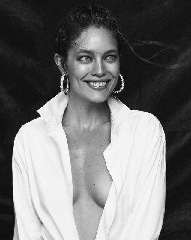 Portrait of a smiling woman with damp hair wearing a white collared shirt and hoop earrings against a dark background