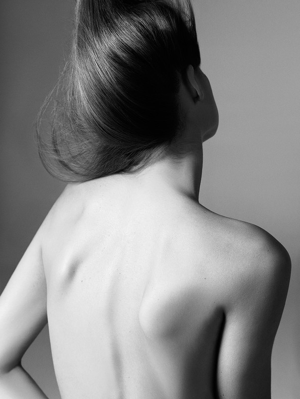 Monochrome back view of a shirtless adult woman with long brown hair up in a studio setting, showcasing the nape of her neck.