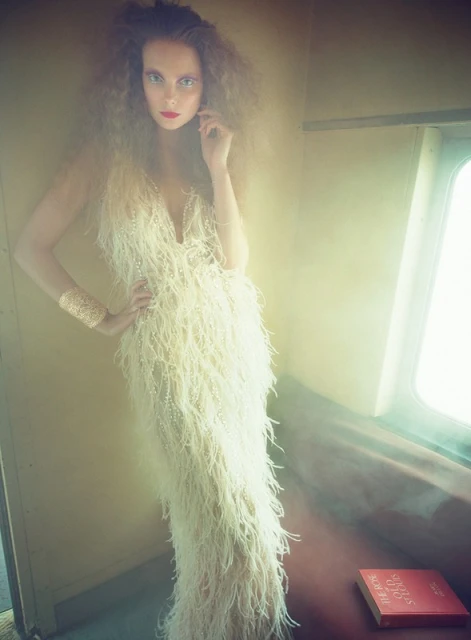 Woman with dramatic makeup poses in feather dress and gold cuff, standing near window and book in indoor fashion editorial setting.
