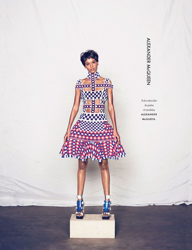 Fashion shot of a Black woman in a checkerboard dress, standing on a block in front of a textured white backdrop in a photo studio.
