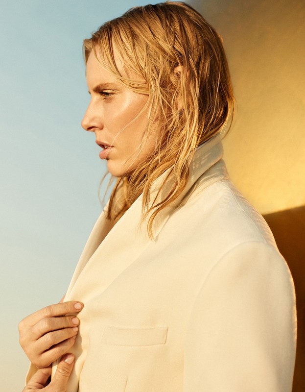 Profile portrait of an adult female with wet blond hair wearing a cream suit jacket, against a color-blocked blue and yellow background.