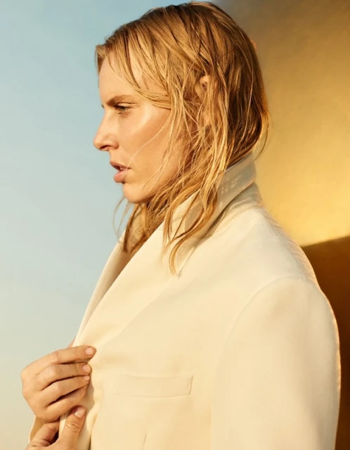 Profile portrait of an adult female with wet blond hair wearing a cream suit jacket, against a color-blocked blue and yellow background.