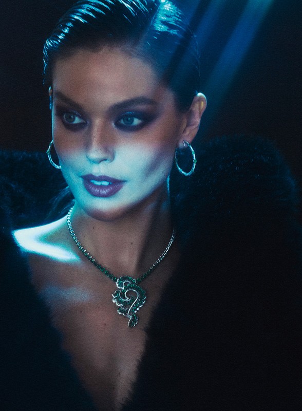 Close-up of a woman with dark hair, hoop earrings, and a pendant necklace, wearing a dark fur stole, lit by blue-toned lighting.