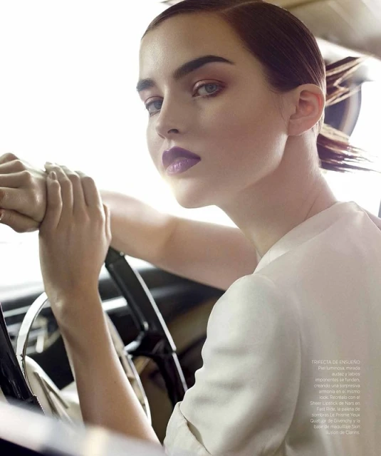Close-up fashion shot of a woman with dark lipstick holding a car steering wheel, styled with slicked-back hair and wearing a white shirt.