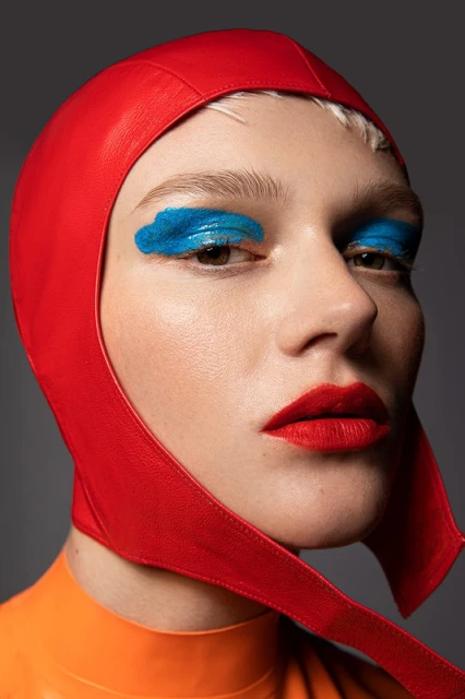 Close-up portrait of a person with blue eyeshadow, red lipstick, wearing a red hood and orange top against a gray background.