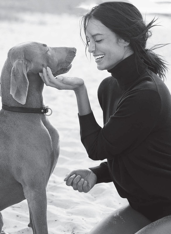 Monochrome shot of a smiling woman kneeling and facing a dog outdoors on a sandy surface in daylight, wearing a dark turtleneck sweater.