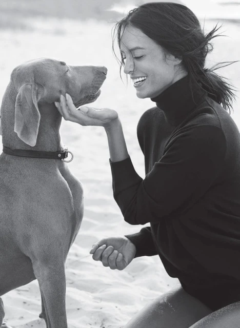 Monochrome shot of a smiling woman kneeling and facing a dog outdoors on a sandy surface in daylight, wearing a dark turtleneck sweater.