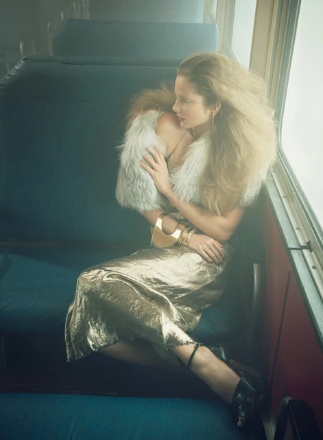 Blonde woman in fur stole and gold dress sits on vintage train seat, looking out the window with soft, diffused lighting.