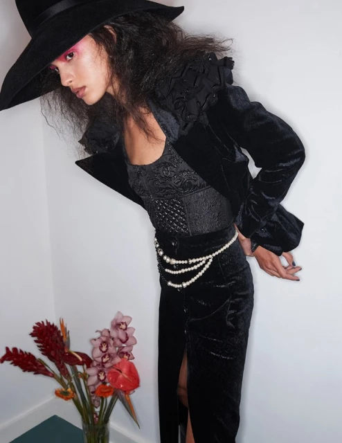 Elegant woman in a black velvet suit and wide-brimmed hat poses indoors with floral arrangement, under soft light, showcasing a high-fashion look.