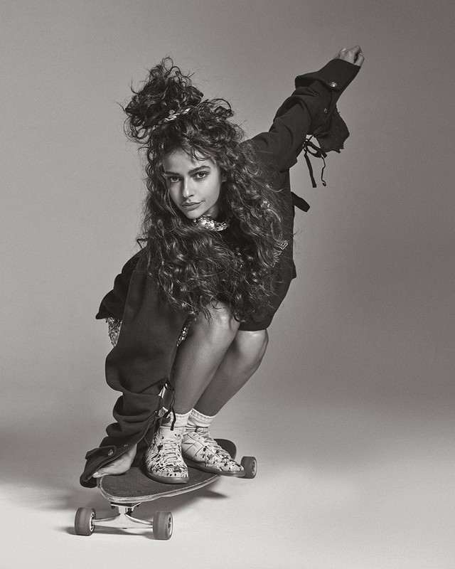 Monochrome portrait of a woman with curly hair squatting on a skateboard against a grey background.