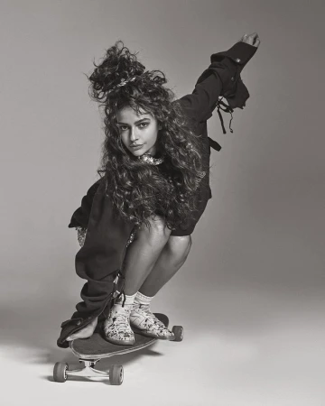 Monochrome portrait of a woman with curly hair squatting on a skateboard against a grey background.