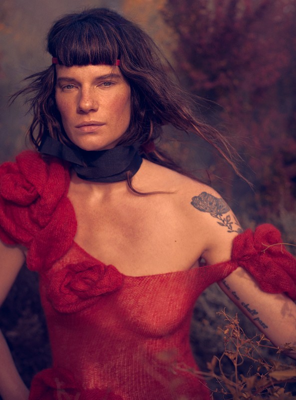 Woman in artistic portrait with red floral shoulder piece, against a dark, moody backdrop. Dark hair frames her face in soft, diffused lighting.