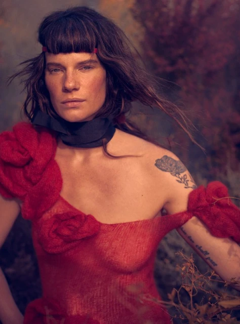 Woman in artistic portrait with red floral shoulder piece, against a dark, moody backdrop. Dark hair frames her face in soft, diffused lighting.