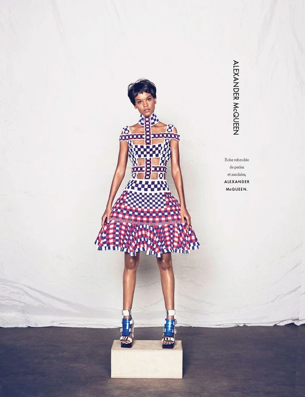 Model with short dark hair wearing a checkered lattice dress standing on a white pedestal against a textured backdrop