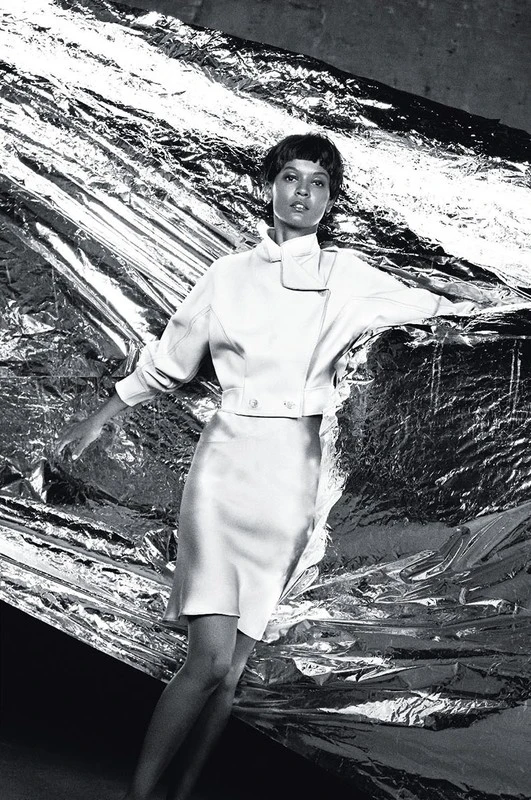 Model with short dark hair in a white jacket and skirt posing against a shiny reflective foil background