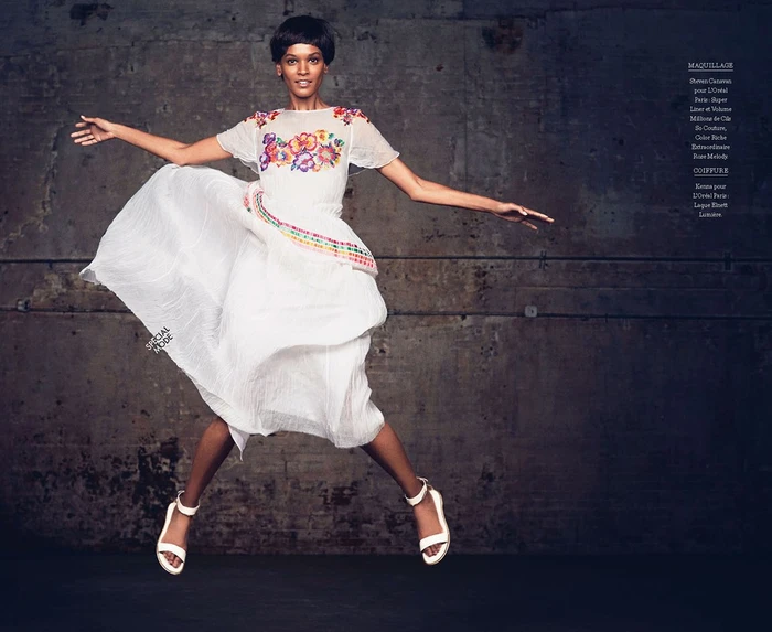 Model in mid-air jumping while wearing a white embroidered dress and white sandals against a textured concrete wall
