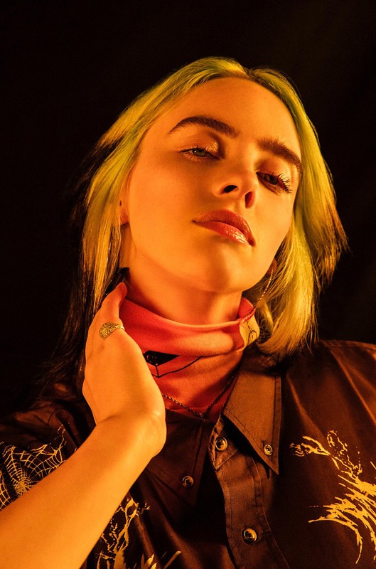Close-up portrait of a young adult female with blond and green hair, wearing a dark patterned shirt in warm, studio lighting.