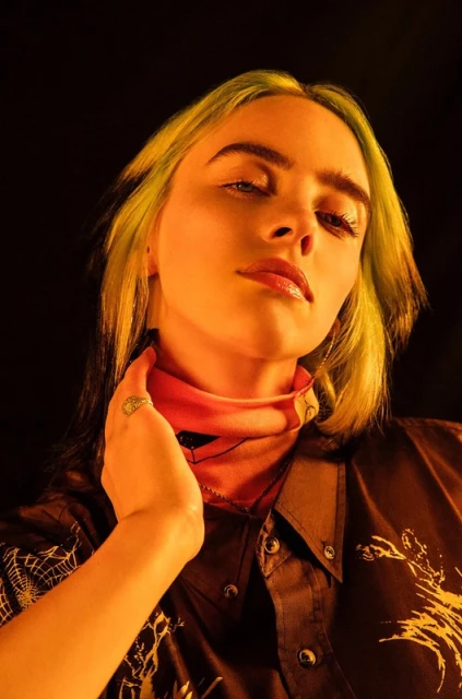 Close-up portrait of a young adult female with blond and green hair, wearing a dark patterned shirt in warm, studio lighting.