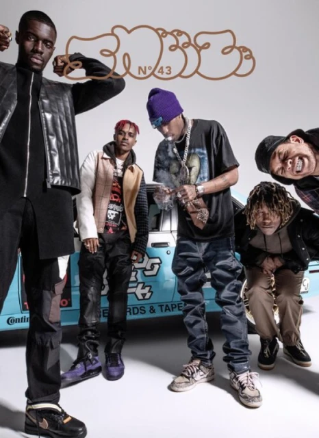 Group of five young adult men in urban fashion standing closely together in front of car with logo on a white background.