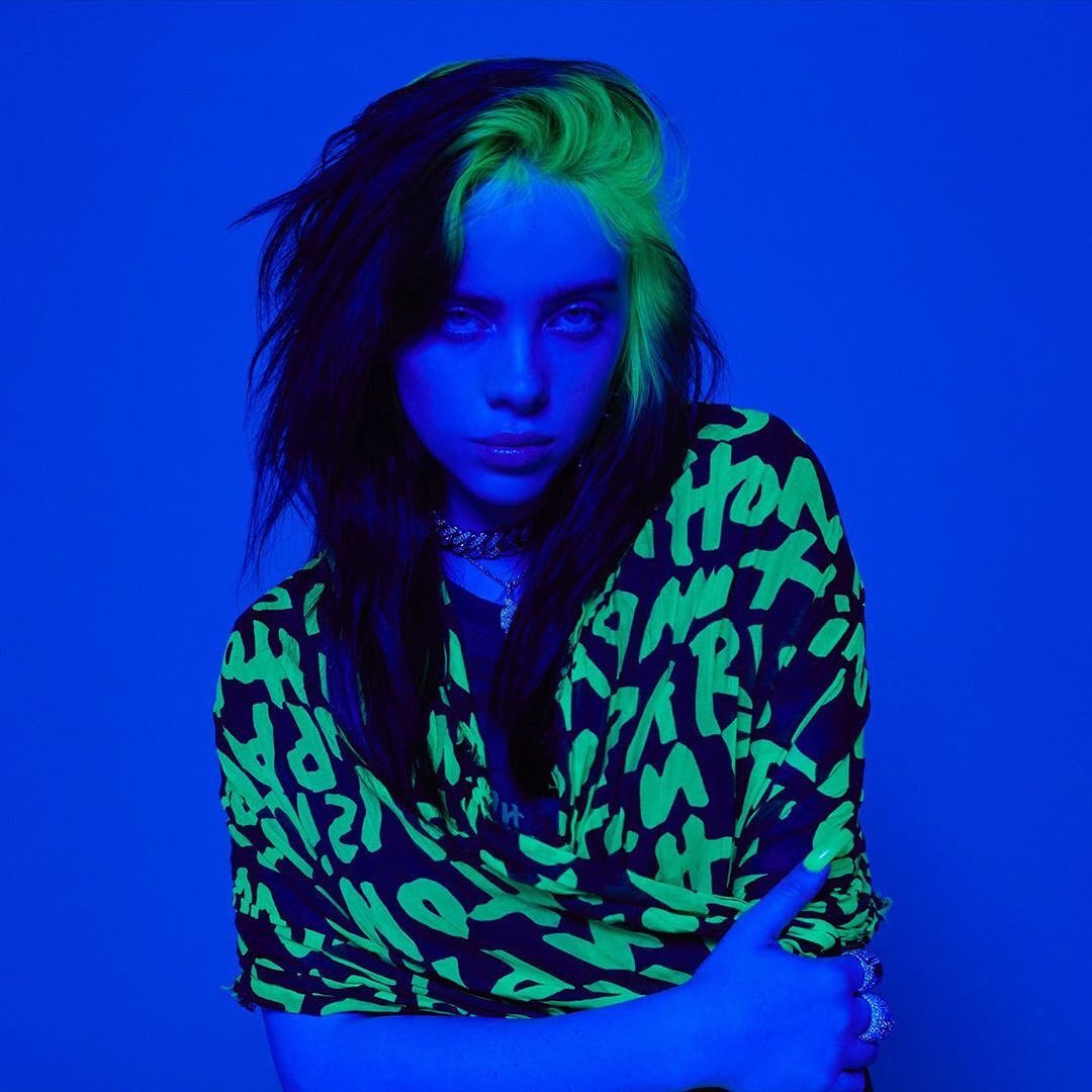 Portrait of adult female with black and neon green hair and green text t-shirt in a blue indoor environment under blue lighting.