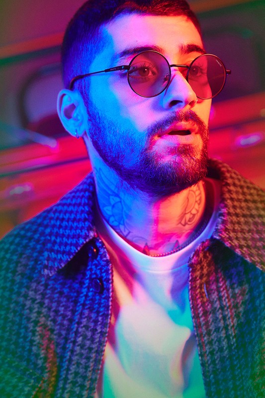 Close-up portrait of adult male with a beard and round sunglasses wearing a patterned coat in a brightly lit indoor setting.