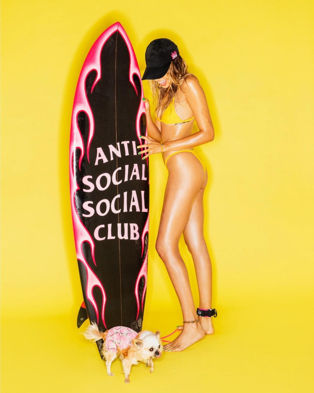 Young woman in a yellow bikini stands next to a surfboard with a dog on a yellow background in a brightly lit photo studio.