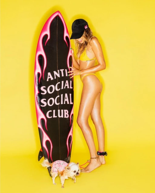 Young woman in a yellow bikini stands next to a surfboard with a dog on a yellow background in a brightly lit photo studio.