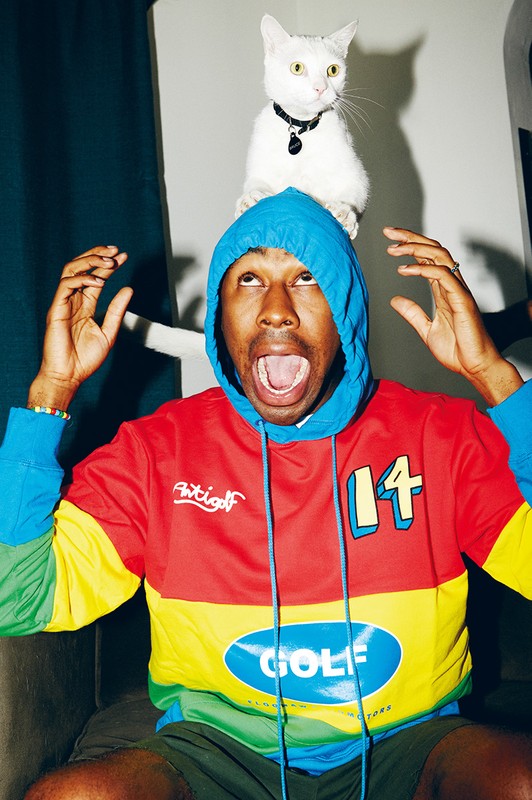Tyler the Creator with a white cat on his head, wearing a colorful hoodie, mouth open, looking upward indoors.