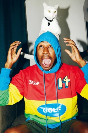 Tyler the Creator with a white cat on his head, wearing a colorful hoodie, mouth open, looking upward indoors.