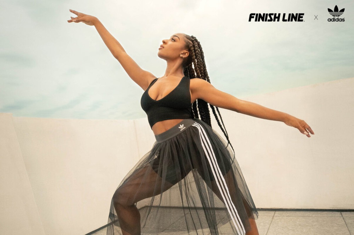 Athletic woman in black sports bra and sheer skirt strikes a pose with arms outstretched against a bright outdoor background.