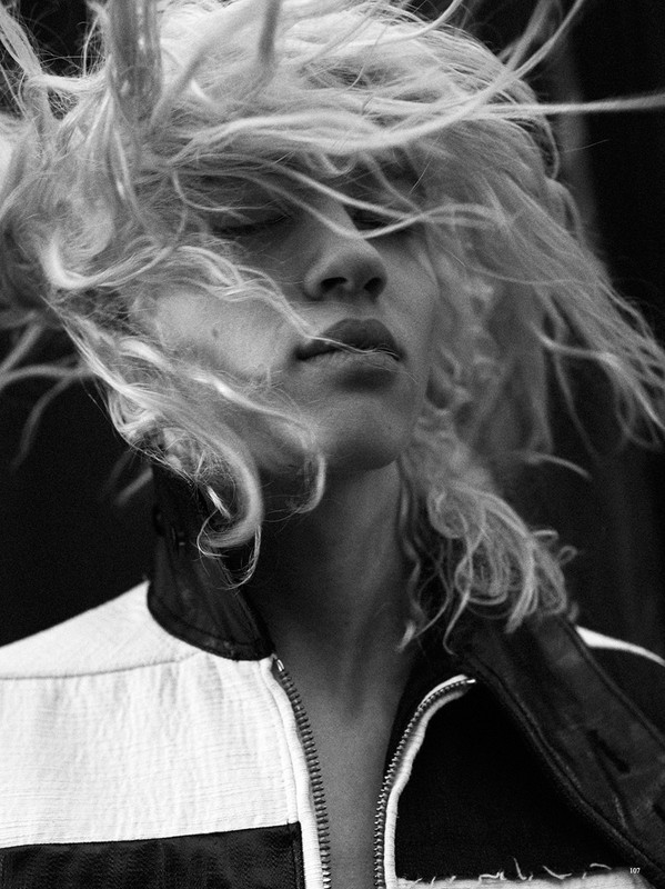 Black and white portrait of a young adult woman with blonde hair blowing around her face, wearing a black and white jacket.