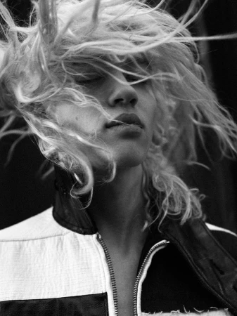 Black and white portrait of a young adult woman with blonde hair blowing around her face, wearing a black and white jacket.