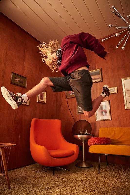 Young adult male mid-air indoors, wearing a red sweatshirt and shorts, curly blond hair, headphones, jumping in a wood-paneled room.