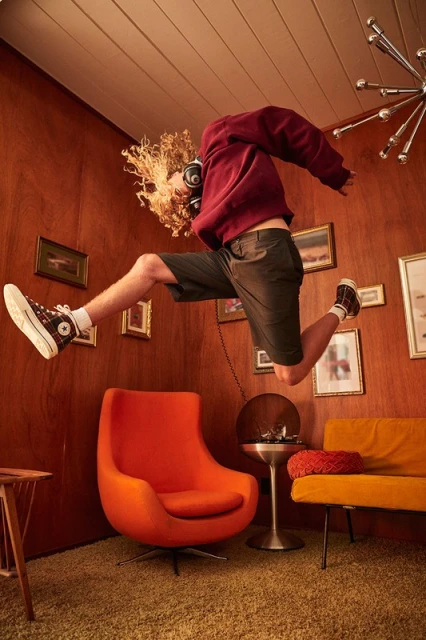 Young adult male mid-air indoors, wearing a red sweatshirt and shorts, curly blond hair, headphones, jumping in a wood-paneled room.