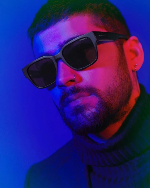Adult male with beard poses wearing black sunglasses against blue and red background; studio lighting creates a modern, fashionable aesthetic.