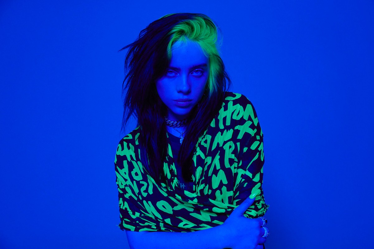 Portrait of Billie Eilish with green-streaked dark hair, wearing a black and neon green patterned top, against a solid blue background.