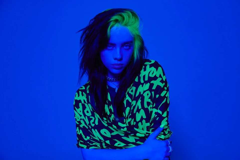 Portrait of Billie Eilish with green-streaked dark hair, wearing a black and neon green patterned top, against a solid blue background.