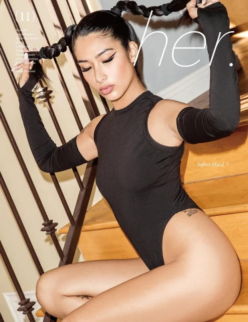 Young woman with braided pigtails poses on wooden stairs wearing a black bodysuit with long fingerless gloves in a studio setting.