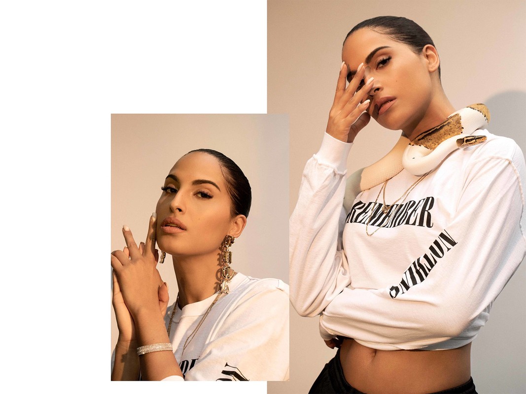 Woman with snake draped on her shoulders poses in photo studio wearing a white long sleeve top, gold jewelry, and black pants.