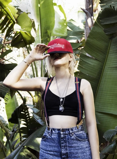 Young adult woman in a baseball cap poses among lush, broad-leafed green plants in natural light. She wears 90s style clothing.