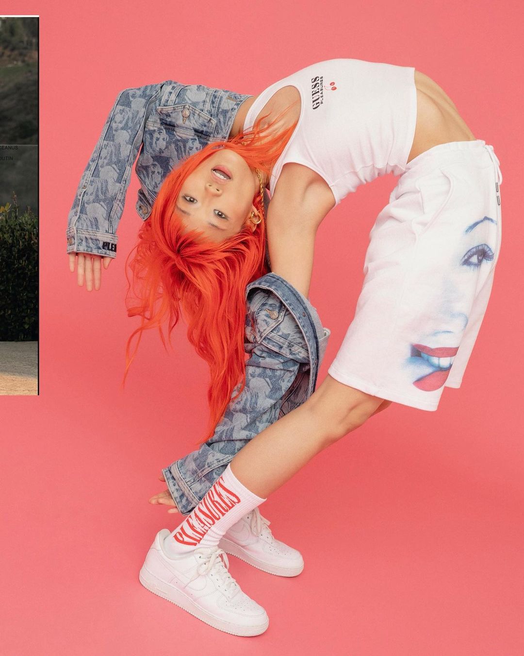 Woman in Gen Z fashion with orange hair strikes a backbend pose against a bright pink background, showing a GUESS top and graphic shorts.