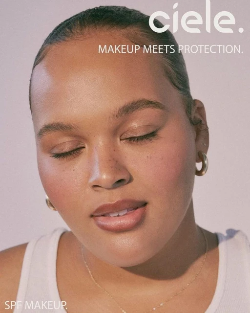 Close-up of a woman with her eyes closed, light makeup, gold hoop earrings, and a gold necklace against a light backdrop. Beauty shot.