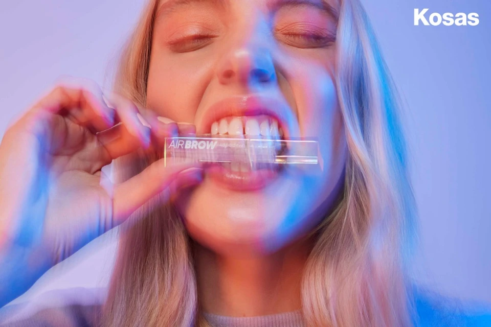 A close-up product shot features a blonde adult female with her eyes closed, holding an Air Brow product by Kosas in her mouth.