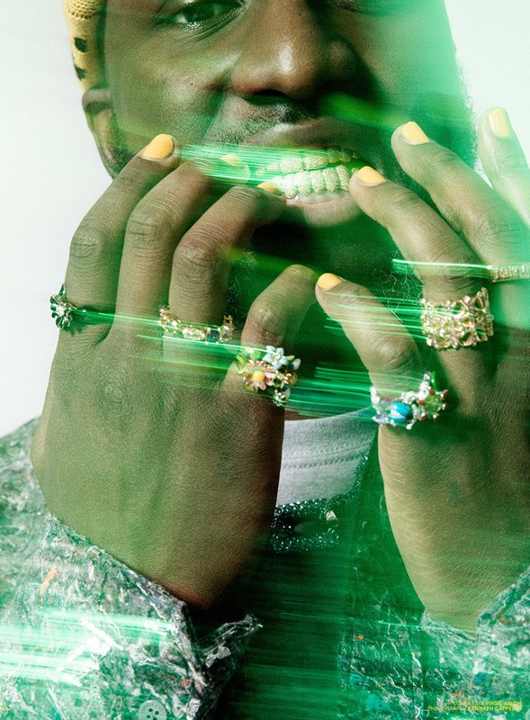Close-up of a smiling Black man with multiple rings on his fingers, wearing a colorful shirt and yellow-green light effects.