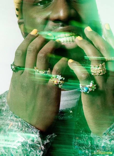 Close-up of a smiling Black man with multiple rings on his fingers, wearing a colorful shirt and yellow-green light effects.