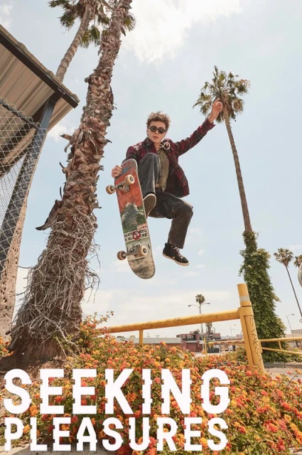 Young man leaps on skateboard in urban setting with palm trees and flowering bushes under bright daylight, showcasing urban action.