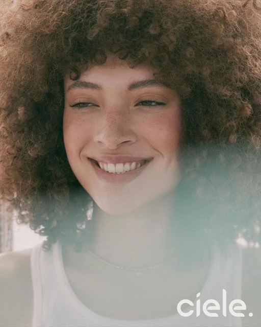 Close-up shot of a smiling woman with curly brown hair and a white tank top indoors, with natural lighting and the brand name 'ciele.'